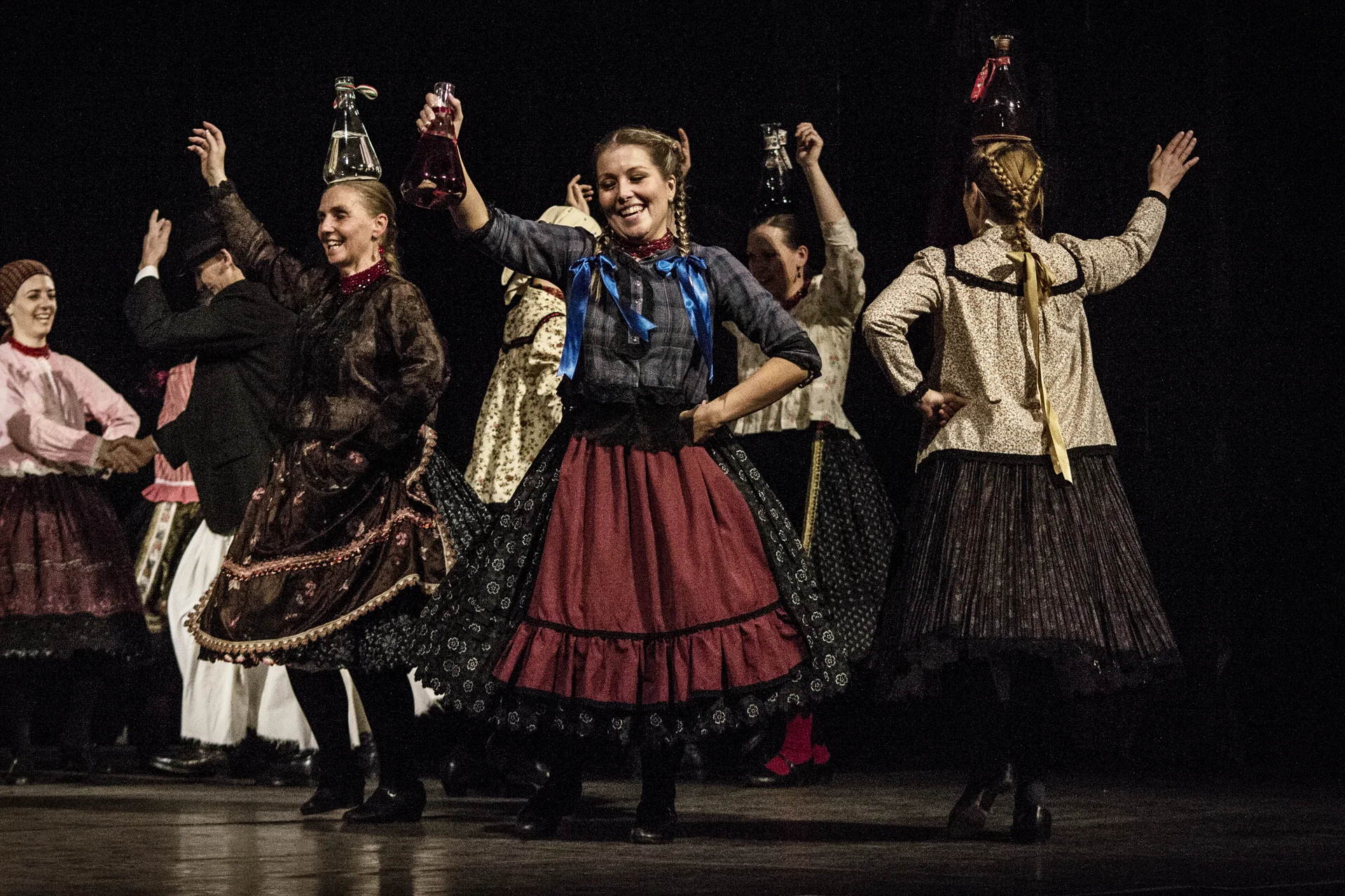 Women in traditional Hungarian folk costume balancing bottles on their heads mid-performance in the bottle dance, on a dark theatre stage